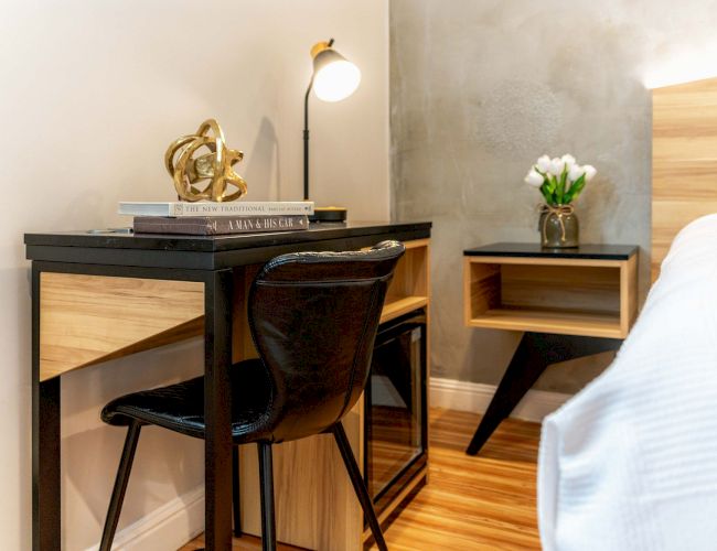 A tidy desk area beside a bed: a small black desk with a chair, books, a lamp, a decorative sculpture, and a nightstand with a plant on it.