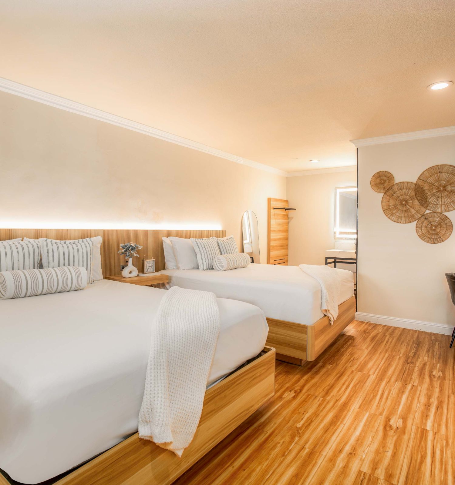 A bright hotel room with two queen beds, wooden floors, soft lighting, a desk with a chair, wall art, and a flat-screen TV, airy and modern.