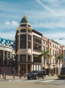 A busy street scene with stylish buildings, palm trees, pedestrians, and a car at an urban corner, likely a shopper-friendly downtown area.