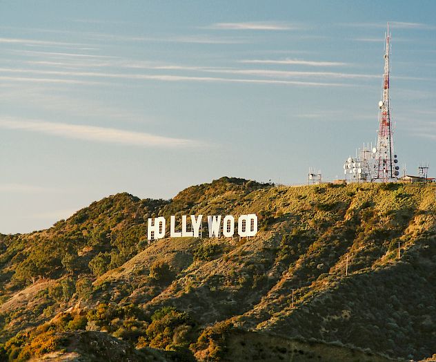 A hillside view of the iconic Hollywood sign in white letters, with a radio tower on the right and clear blue sky above.