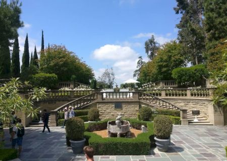 A formal garden with stone steps, trimmed hedges, and a central seating area; people stroll the sunlit terrace under a blue sky.