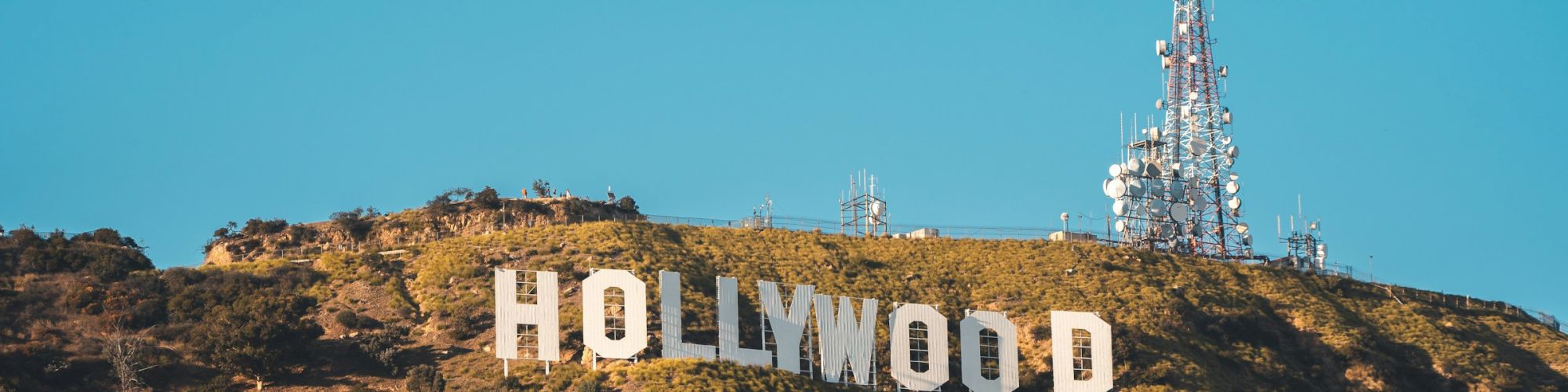 A hillside with classic Hollywood sign letters and a large antenna/tower on the peak against a clear blue sky.
