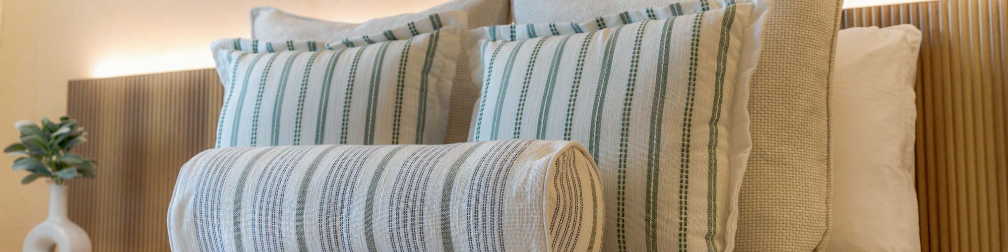 A neatly made bed with striped pillows and a cylindrical bolster, soft lighting, and a small plant on the bedside table, calming hotel-room vibes.