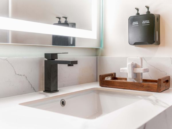 A modern bathroom sink setup with a black matte faucet, white countertop, backlit mirror, and a wooden tray holding white towels on the right.