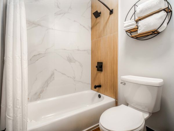 A compact bathroom with a white tub, marble-look walls, wooden accent panel, towel hooks, a wall basket, and a white toilet. This room is bright and modern.