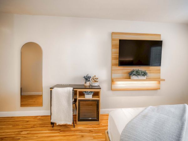 A modern hotel-style room with a mounted TV above a wooden shelf, a small kitchenette area with a mini-fridge, and a bed with light bedding, plus a decorative mirror alcove in the wall, all on warm wood flooring.