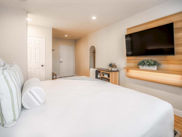 A cozy hotel-style bedroom with a mounted TV on a wooden panel, a white bed with striped pillows, and a small desk area by the door, end of sentence.