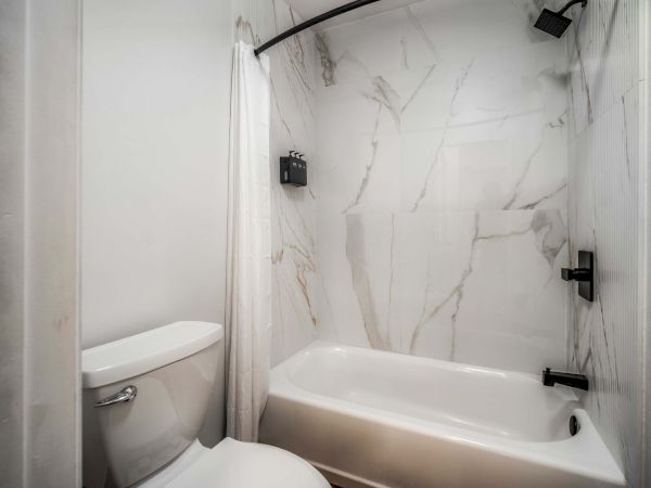A small bathroom with a white toilet and a marble-tiled shower/bath combo, black fixtures, and a curved shower curtain rod.