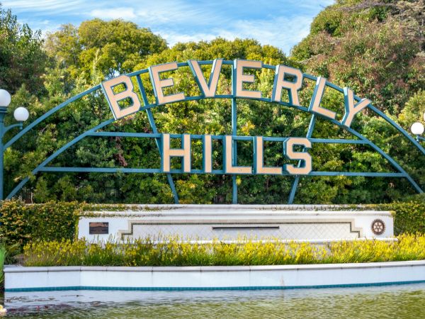 The image shows the iconic Beverly Hills sign surrounded by greenery and lamps, with a small body of water in front of it.