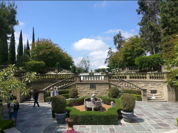 The image shows an outdoor garden with stone staircases, well-manicured greenery, and a central fountain under a partly cloudy sky.