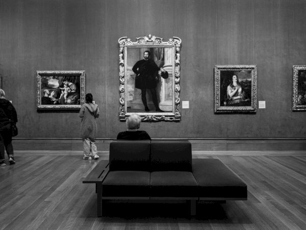 People observe artworks in a gallery, with paintings framed on the wall and one person seated on a bench in the center of the room.