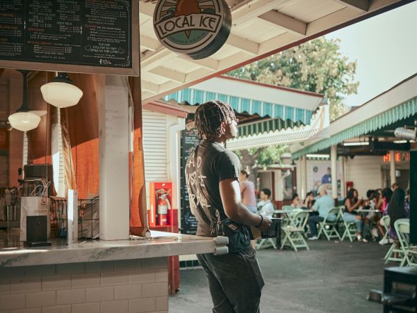 A person stands at an outdoor food stand, looking up at a sign. People are seated at tables in the background. The setting appears casual.