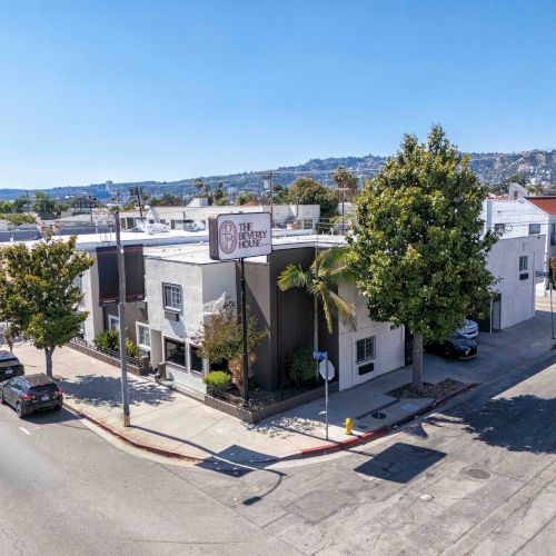 A small white building on a corner, street lined with parked cars, trees, and a clear blue sky over a quiet urban block.