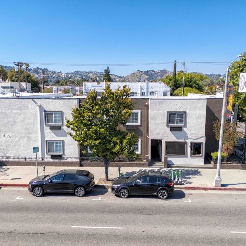 A street view of a row of light-gray two-story buildings with parked cars, a tree in front, and a clear blue sky above.