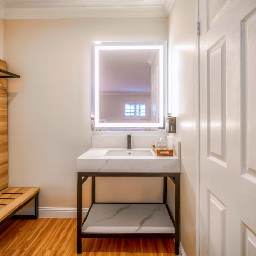 A small dressing area with a wooden bench and rack, a sink with a lit-framed mirror, and a metal-and-wood table beneath a ready-to-use sink, neat and minimal.