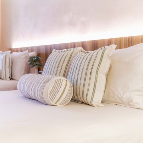 A clean, neatly made hotel bed with white sheets, striped pillows, and a round bolster, soft ambient light in a cozy bedroom.