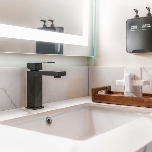 A modern bathroom sink with a black faucet, white marble counter, a wooden tray, and a wall-mounted soap dispenser on the tiled wall.