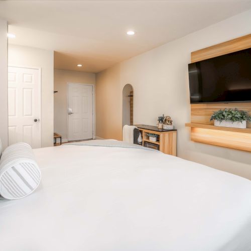 A cozy hotel-style bedroom featuring a plush white bed, striped pillows, wall-mounted flat-screen TV, wooden accent panel, and a small desk area near a doorway.
