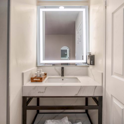 A modern bathroom vanity with a square backlit mirror, white marble sink, and a metal frame beneath; towels neatly stored below.