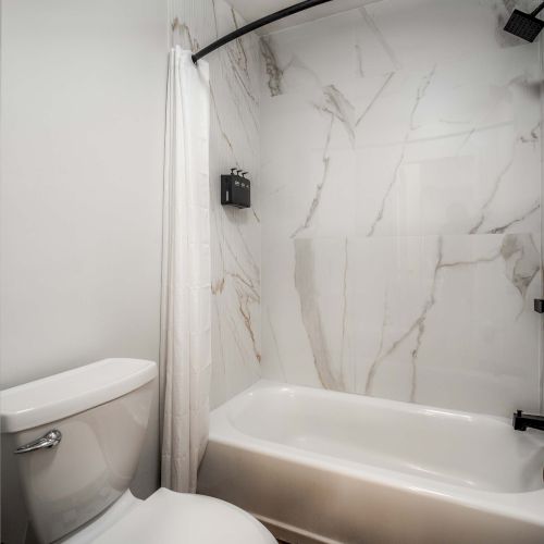 A small bathroom with a toilet and a bathtub/shower combo, marble-patterned walls, a curved shower rod, and dark fixtures.