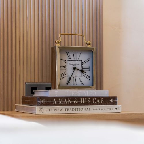A vintage clock sits atop a stack of books on a bedside table, with warm wooden panels in the background.