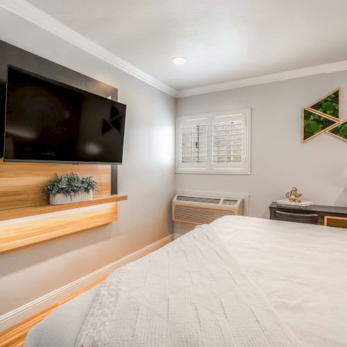 A modern bedroom with a wall-mounted TV and floating wooden console, a cozy bed, a small desk with a lamp, and light gray walls under a window, all tidy and bright.