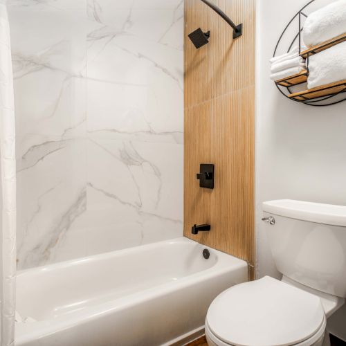 A modern bathroom with a white tub and shower, marble tile wall, wooden accent panel, black fixtures, a white toilet, and a round wall shelf with towels.