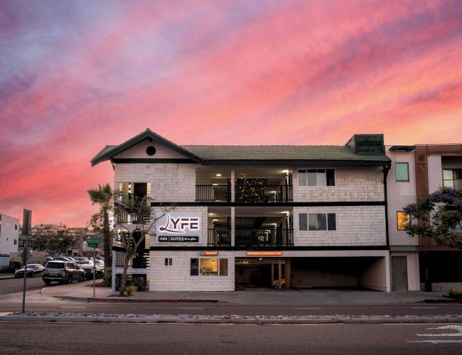 A three-story white brick building with a parking area underneath, a sign &ldquo;YF&rdquo; on the left, and a dramatic pink sunset sky.