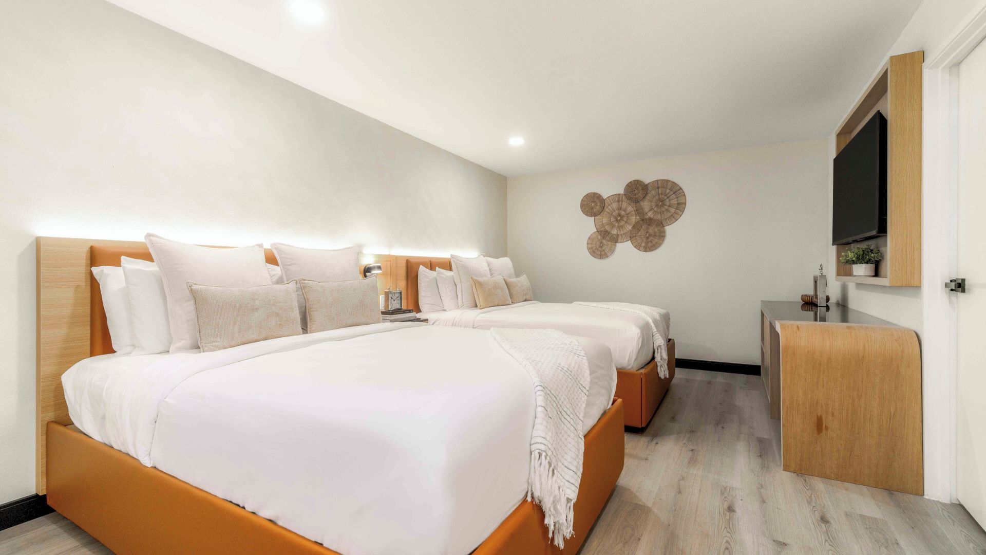 A modern hotel room with two large white beds, soft gray walls, wooden accents, a flat-screen TV, and light hardwood floors.
