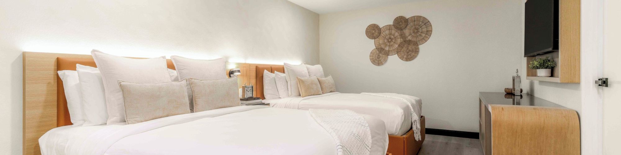 A modern hotel room with two large white beds, soft gray walls, wooden accents, a flat-screen TV, and light hardwood floors.