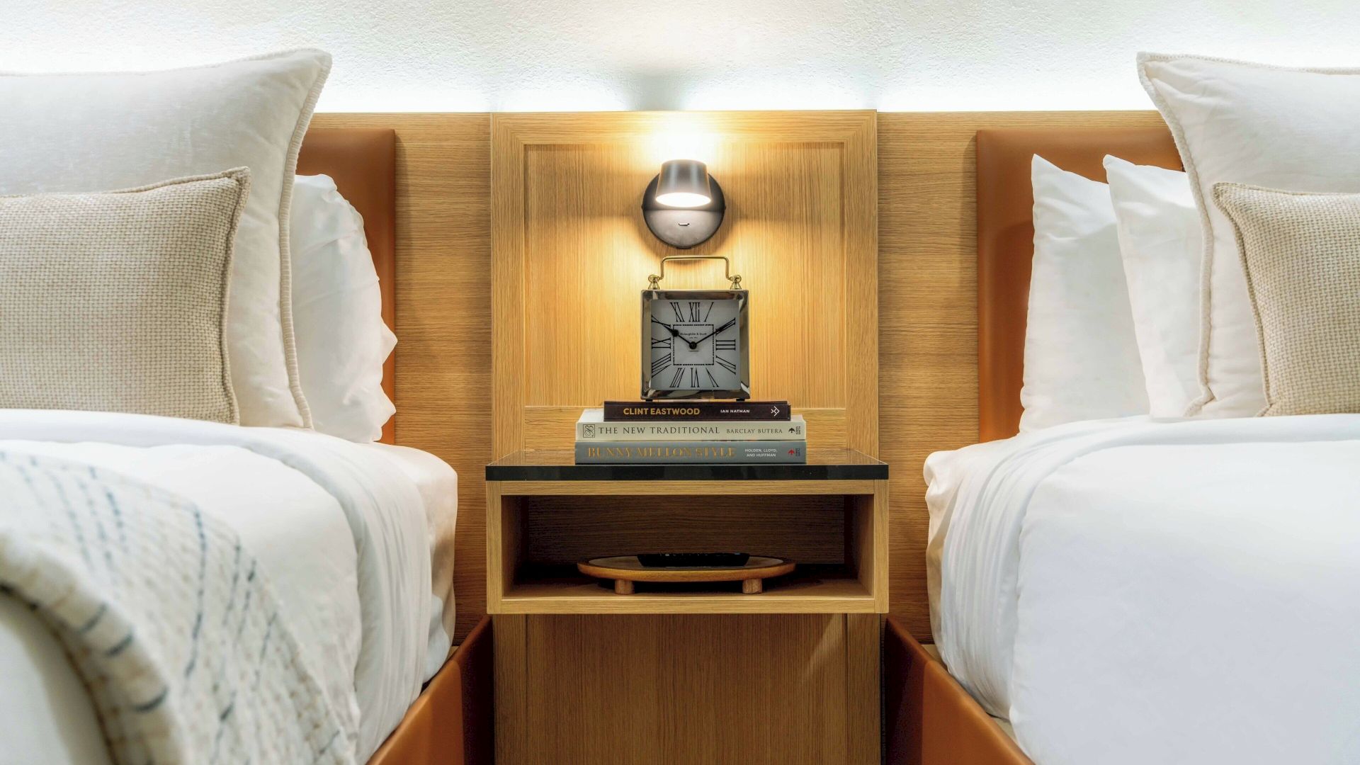 A hotel bedroom scene with two beds, a wood headboard, a bedside lamp, and stacked books on a central nightstand.