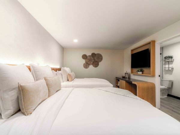 A bright hotel room with two double beds, fluffy white linens and beige pillows, a wall art piece, desk with TV, and a connected bathroom.