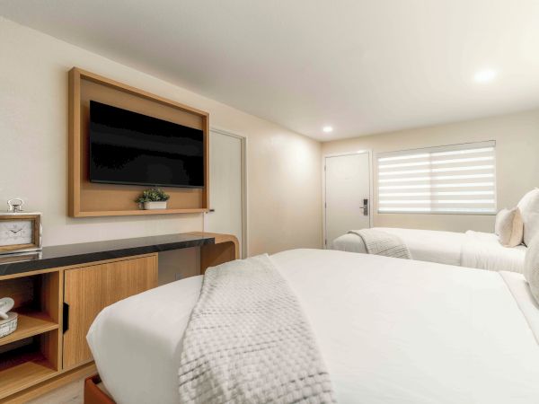 A modern hotel-style bedroom with two single beds, wall-mounted TV, light wood furniture, and a bright, minimalist decor, cozy and clean.