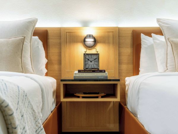 Two neatly made hotel beds with a nightstand between them, a vintage clock on top, and soft lighting above; cozy, symmetrical room.