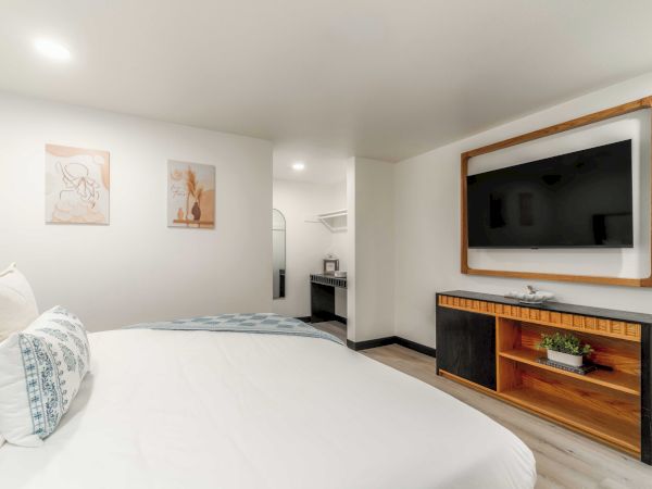 A bright hotel room featuring a large bed with white linens, cushioned pillows, wall art, a wall-mounted TV in a wooden frame, and a console with storage.