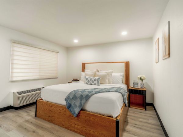 A modern bedroom with a wooden bed, white and gray bedding, a blue throw, two matching nightstands, a window with blinds, and minimalist decor.