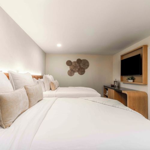 A bright hotel room with two large beds, neutral linens, a wall-mounted TV, a desk, and an open bathroom door at the far end, modern and clean.