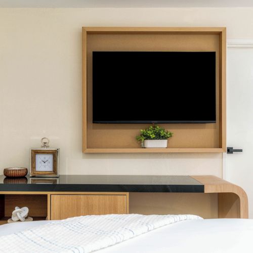 A stylish bedroom with a wall-mounted wooden shelf and TV, a black countertop, small plant, clock, and bedside items, plus a white bed in the foreground.