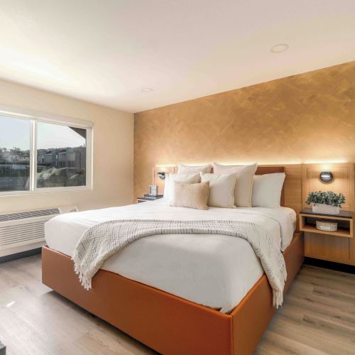 A bright hotel bedroom with a large bed, plush white bedding, soft beige accent wall, two bedside tables, a window view, and minimalist decor.