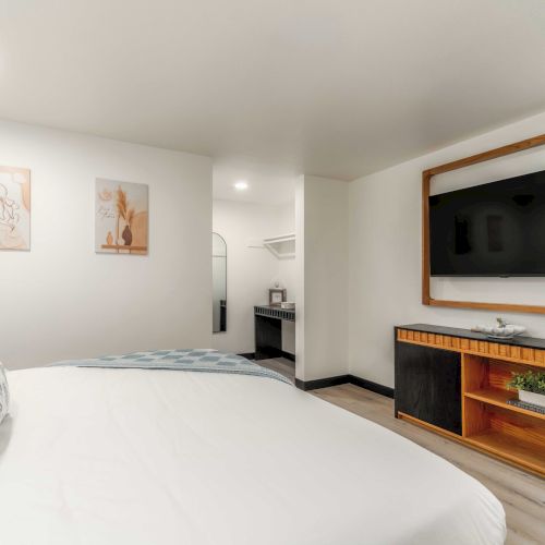 A modern hotel room with a large bed, white bedding, wall art, and a flat-screen TV in a wooden frame, plus a small cabinet and desk area.
