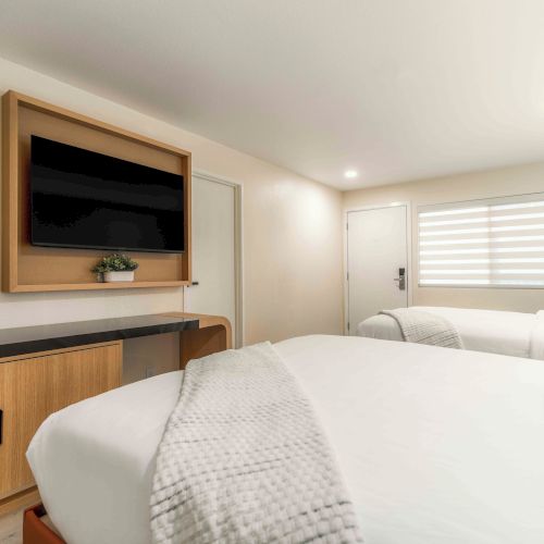 A modern hotel room with two neatly made beds, a wall-mounted TV, desk area, and soft natural light from a window with blinds, minimal decor.
