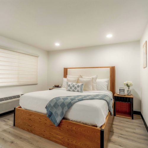 A bright, modern bedroom with a wooden bed frame, white bedding, gray throw, two matching nightstands, and minimal wall decor, plus a window and baseboard heater nearby.