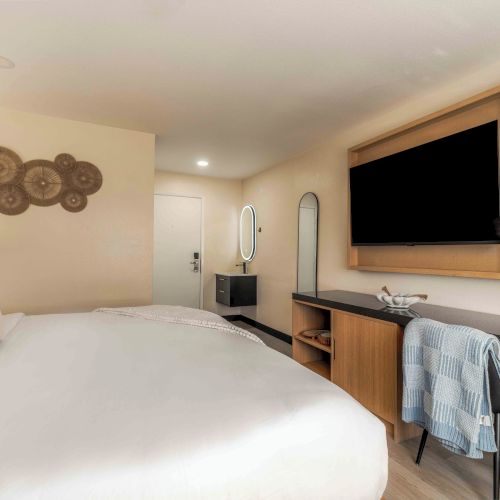 A cozy hotel room with a large bed, a desk and chair, a wall-mounted TV, and a dresser with a round mirror, all in warm, neutral tones.