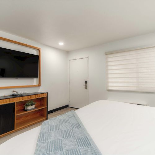 A modern hotel-style bedroom with a wall-mounted TV, a wooden console below, a closed door, a window with blinds, and a neatly made bed with blue accents. (End with a period.)