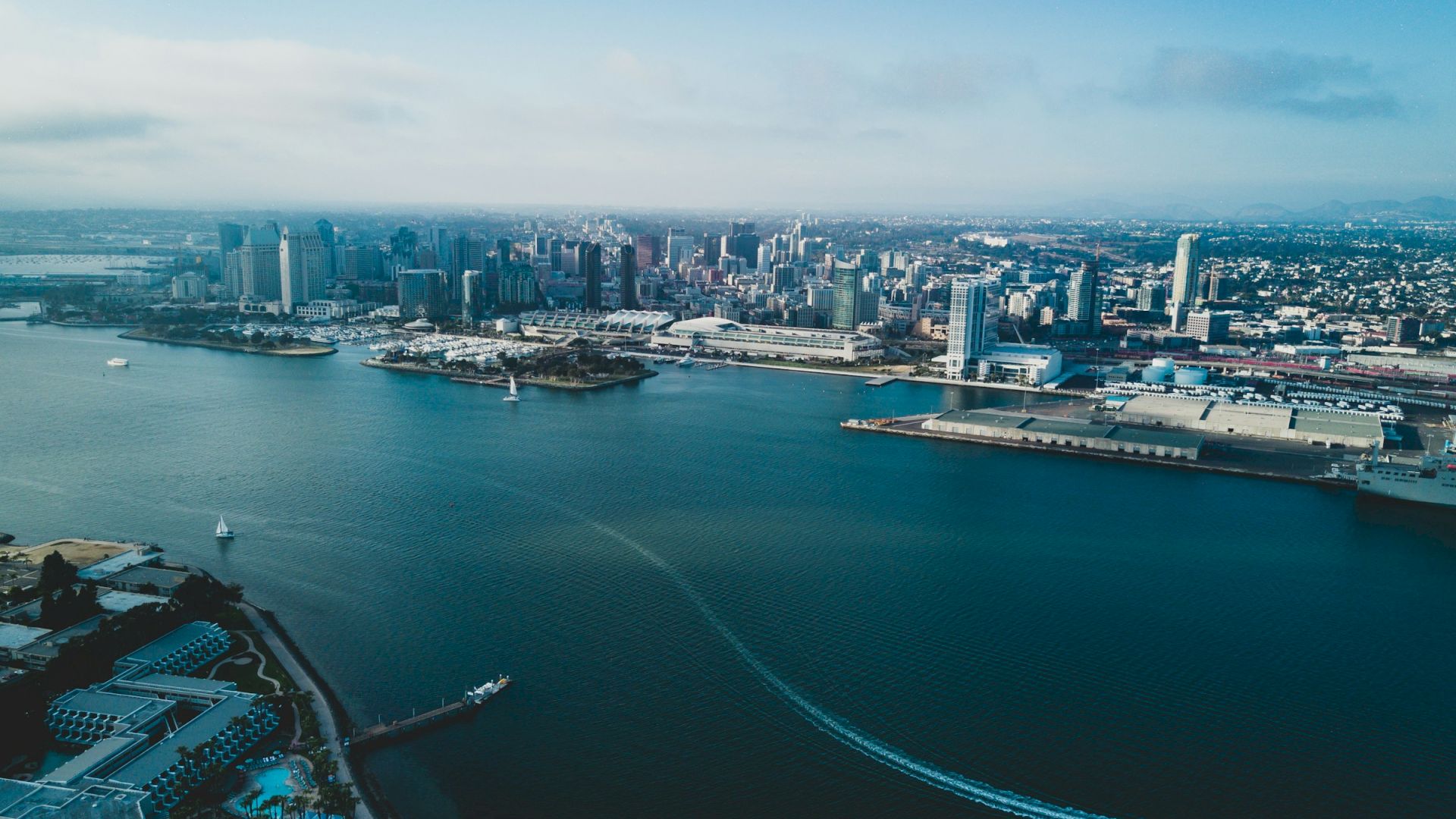 Aerial view of a coastal city with a large harbor, tall buildings in the skyline, and a boat trailing a white wake across calm blue water, ending with the horizon.