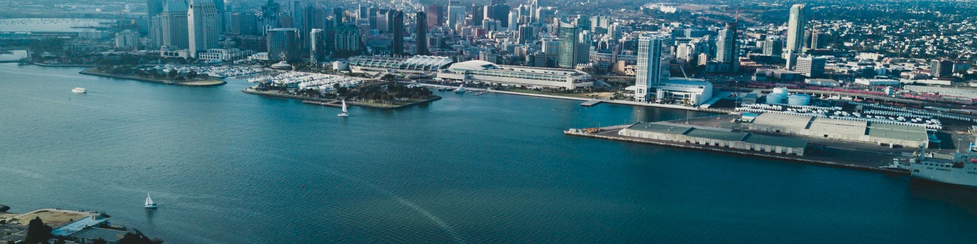 Aerial view of a coastal city with a large harbor, tall buildings in the skyline, and a boat trailing a white wake across calm blue water, ending with the horizon.