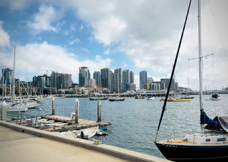 A bright marina with sailboats docked along a calm harbor and a modern city skyline in the background, under a partly cloudy blue sky.