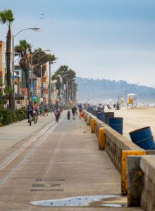 A sunny beachfront boardwalk with cyclists and pedestrians, palm trees, and colorful shops along the coast, near a calm ocean and sandy beach.