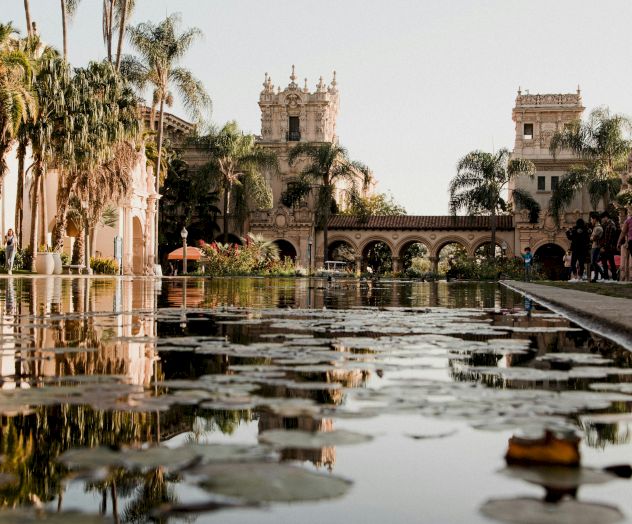 A serene garden scene with palm trees, a reflective pool, and a historic architectural backdrop under a clear sky.