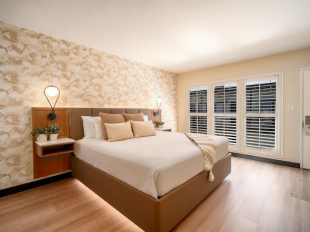 A modern hotel room with a large bed, patterned feature wall, wood nightstands, soft lighting, and white shutters by a glass door.